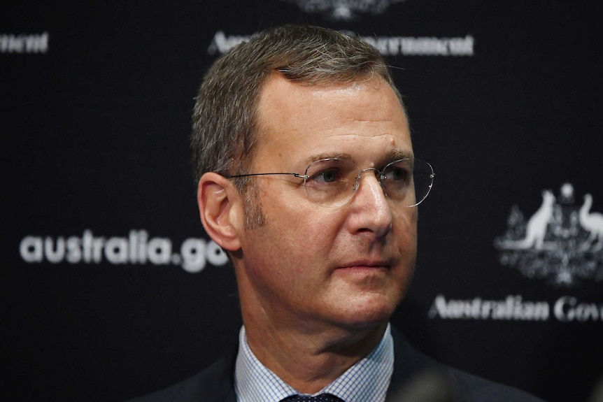 Michael Kidd stands in front of a Government banner and looks to his left.