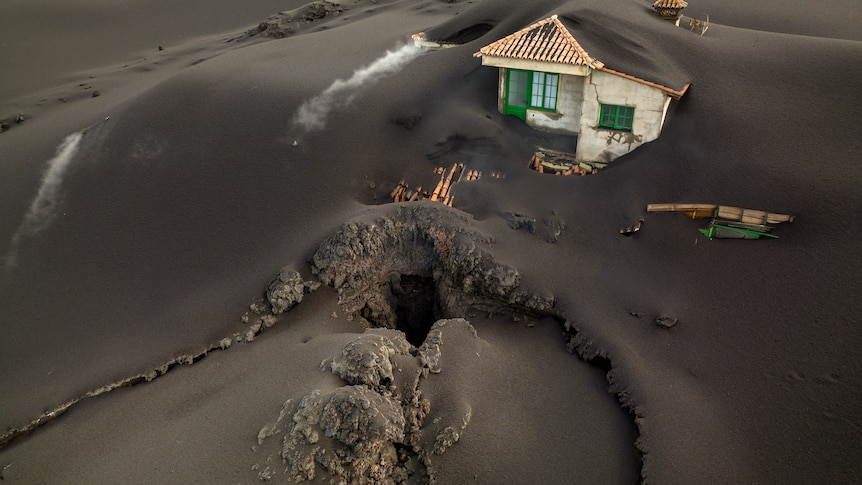 A large hole emerges in the earth in front of a house which is completely covered in grey ash.