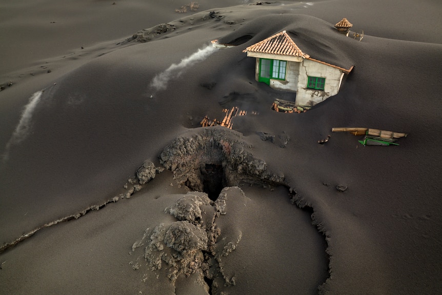 Un gran agujero emerge en la tierra frente a una casa que está completamente cubierta de ceniza gris.