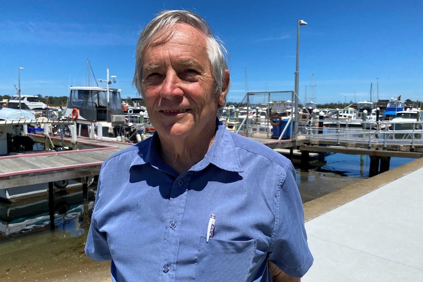 An elderly man standing in front of a jetty with boats in the background