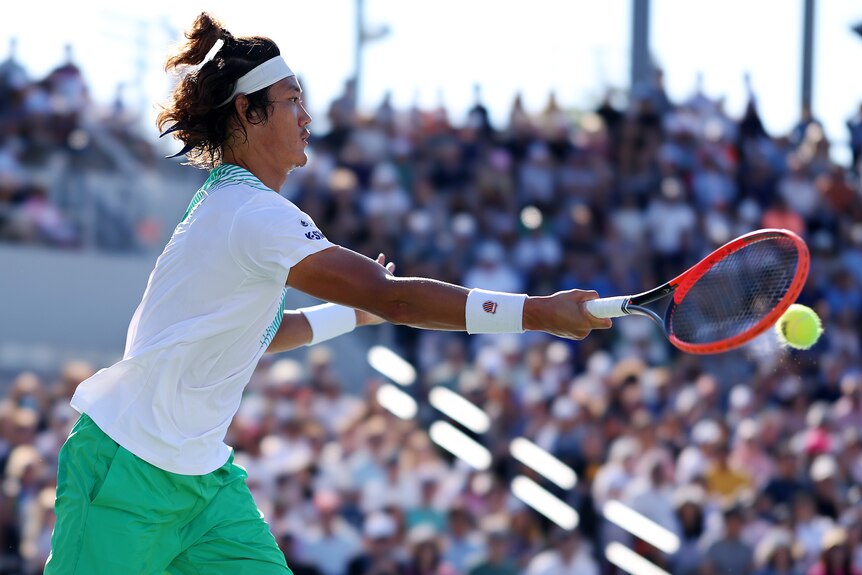 A male Chinese tennis player, wearing a white top and green shorts, plays a forehand