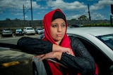 Huma Naz standing next to the car, leaning on the door and looking into the distance.