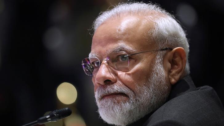 Narendra Modi is seen in a headshot against a dark background. A microphone is in front of him and he is listening.