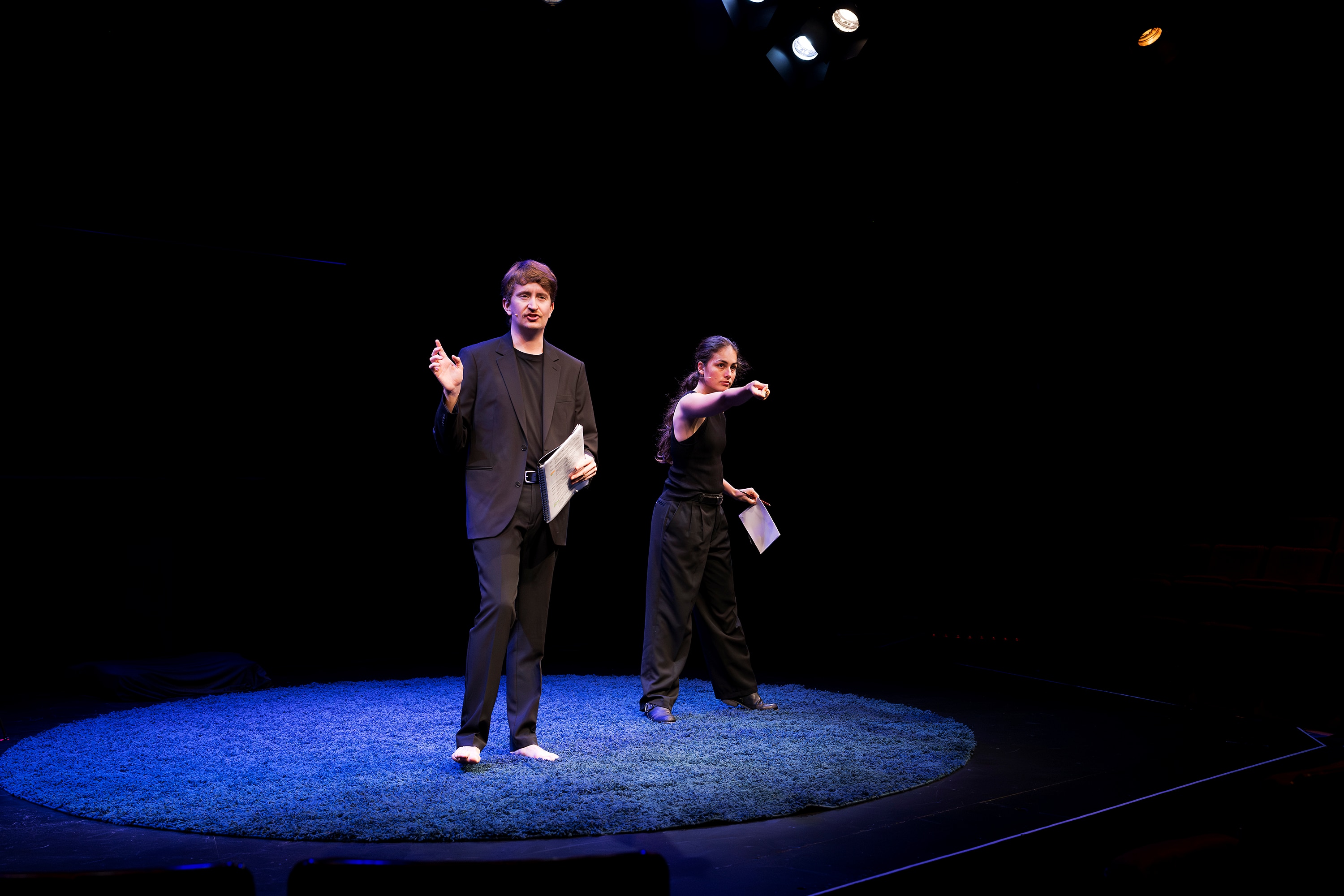 Young man and young woman, both wearing black, on stage under spotlight