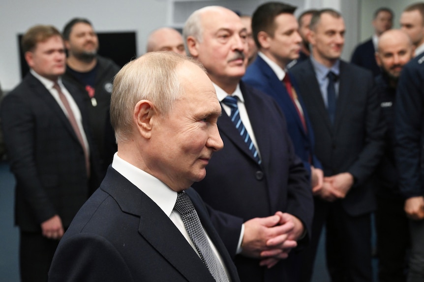 A balding older Russian man in a suit smiles among a crowd of other men in suits.