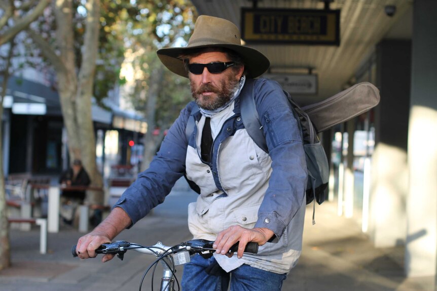 Gary sits on his bike, wearing an akubra hat and carrying what looks like a small stringed instrument on his back.