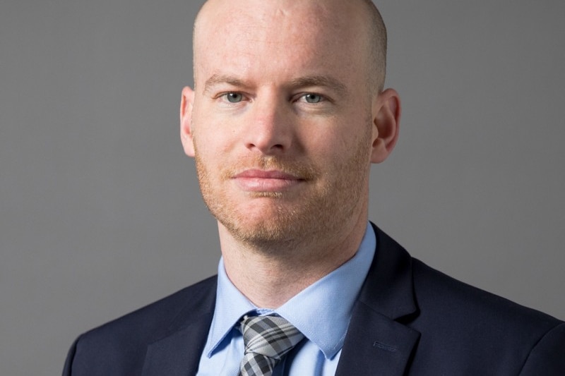 A head shot of slightly smiling man in a blue suit, light blue shirt and check tie.