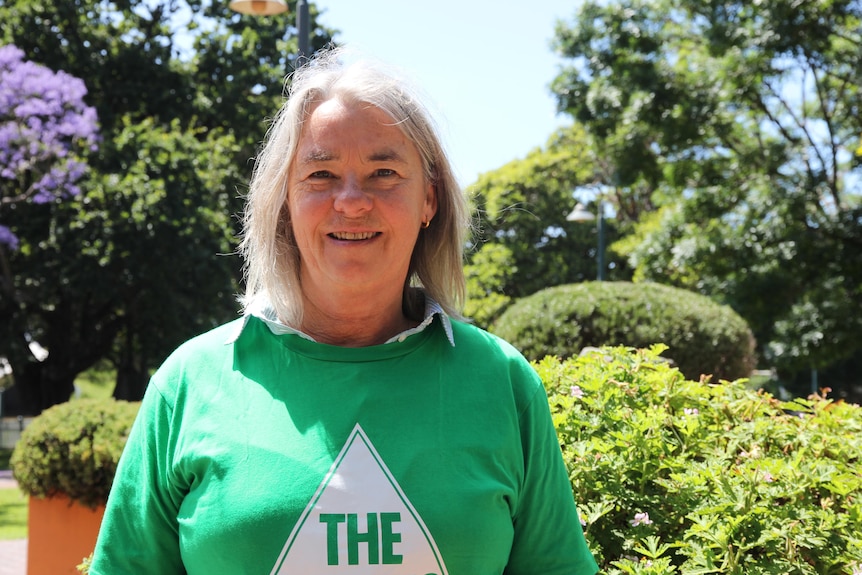 A woman stands with a green 'greens' t-shirt on in front of shrubs