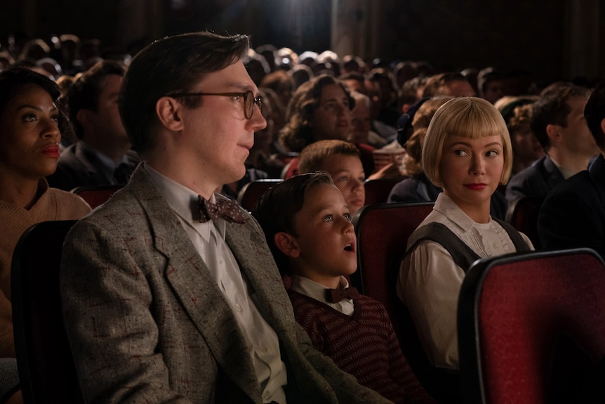 A white boy in vest and bow-tie sits between a white man in tweed jacket and white woman with short blonde bob in cinema seats.