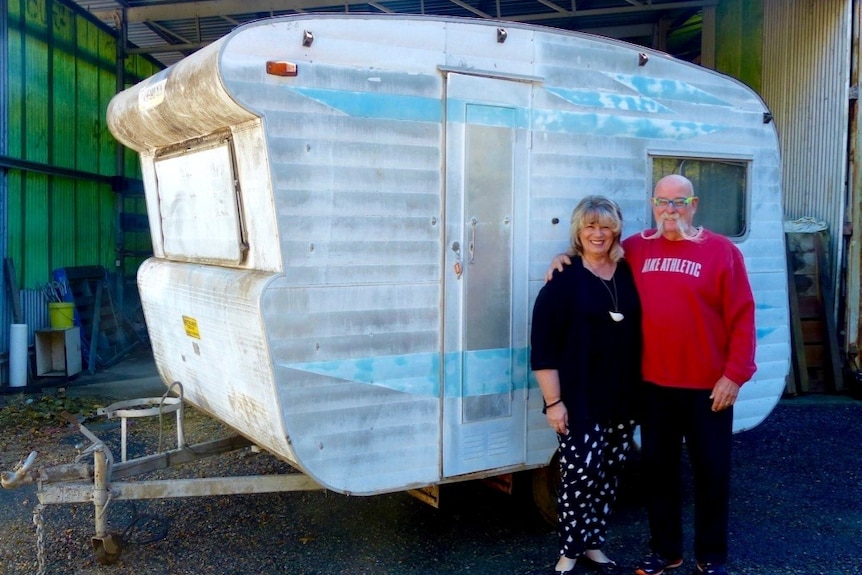 A couple stand smiling in front of an unrenovated caravan.