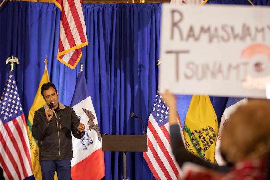 Vivek Ramaswamy holds a microphone and speaks in front of a blue curtain and large flags. A sign says 'Ramaswamy Tsunami'