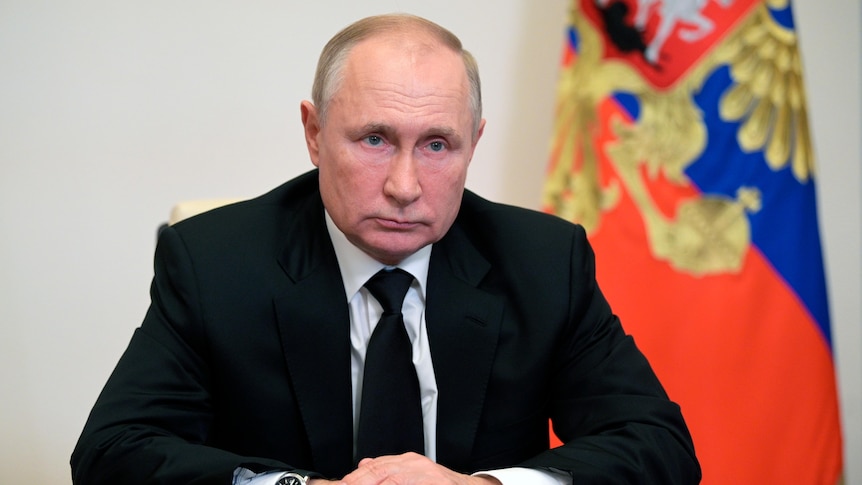 一名穿着黑色西服的男子坐在办公桌边，身后是俄罗斯国旗。