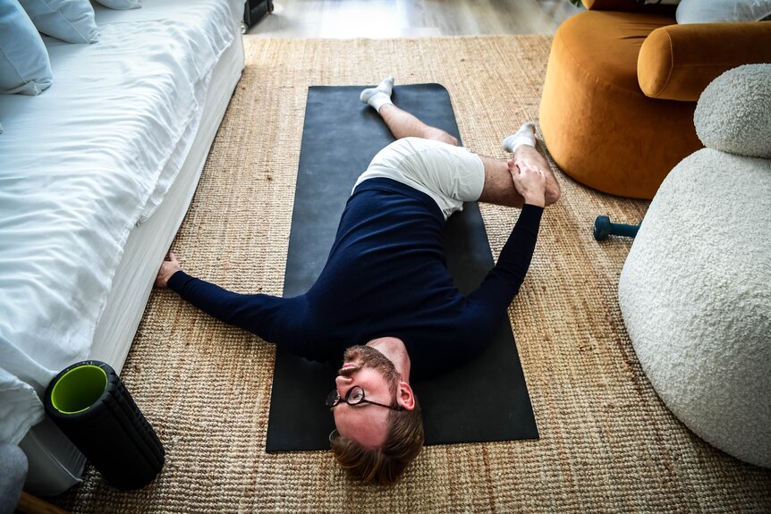 Giles lies on a yoga mat on the floor. He is twisting his body, pulling his knee in one direction while he faces the other.