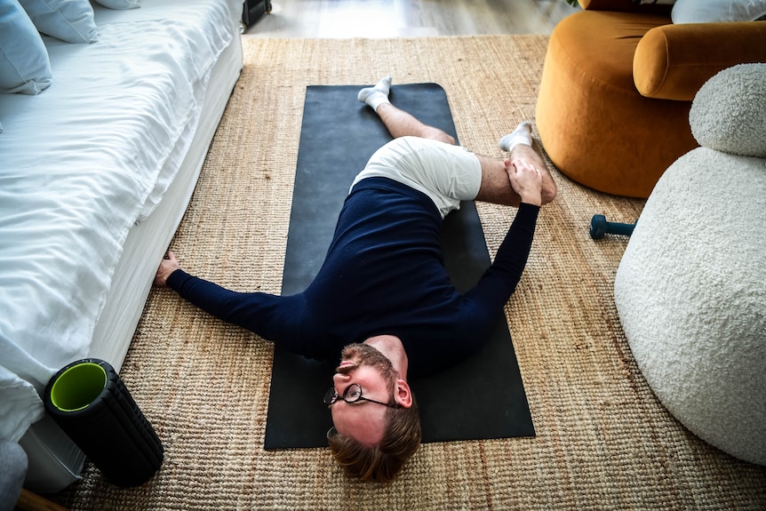 Giles lies on a yoga mat on the floor. He is twisting his body, pulling his knee in one direction while he faces the other.