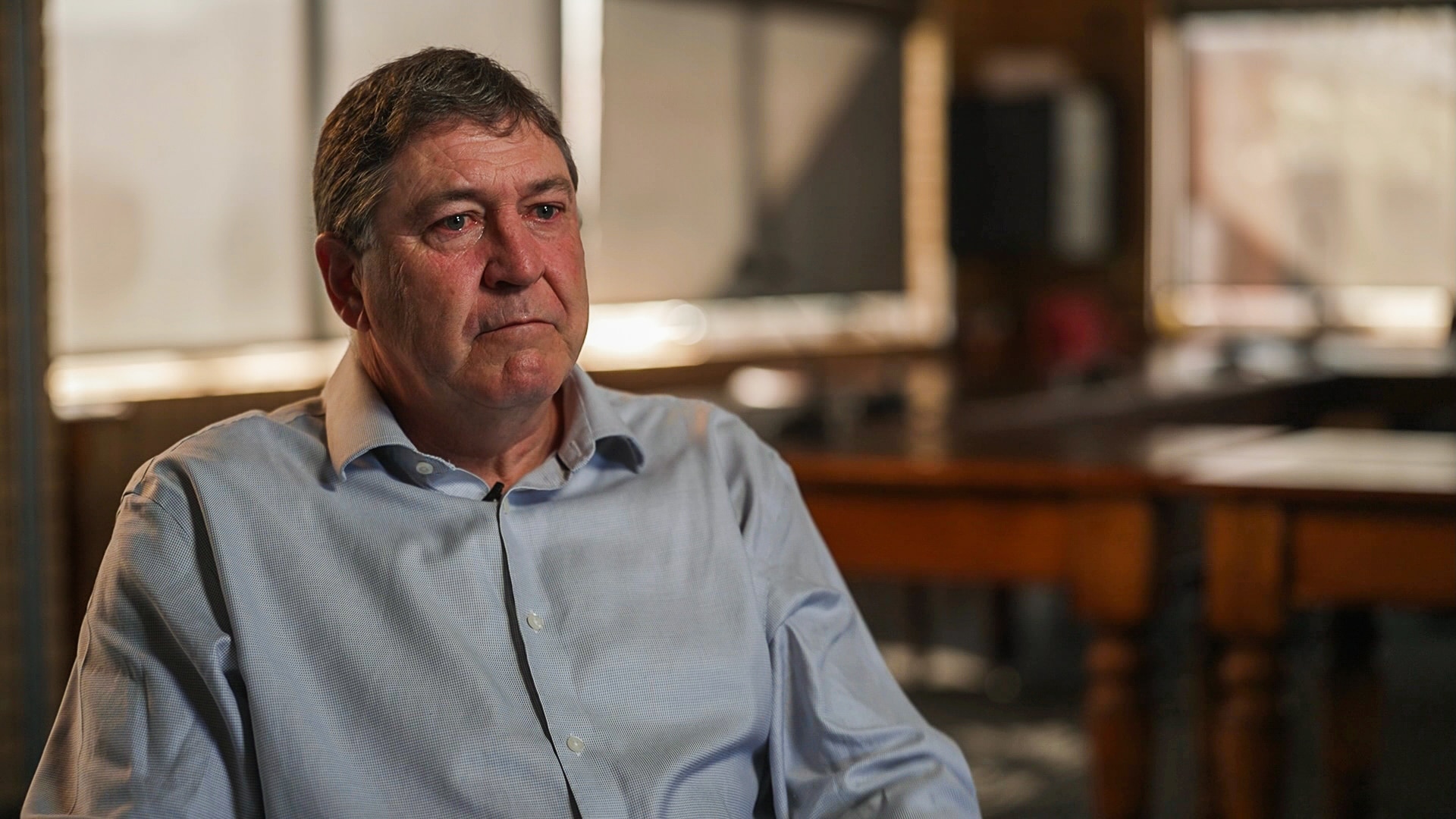 Man in blue button up shirt sits in front of camera. His eyes look teary
