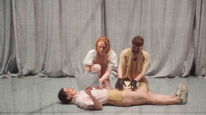 On a stage behind mesh material three dancers, one lies on the ground holding a piece of paper and the others touch her body