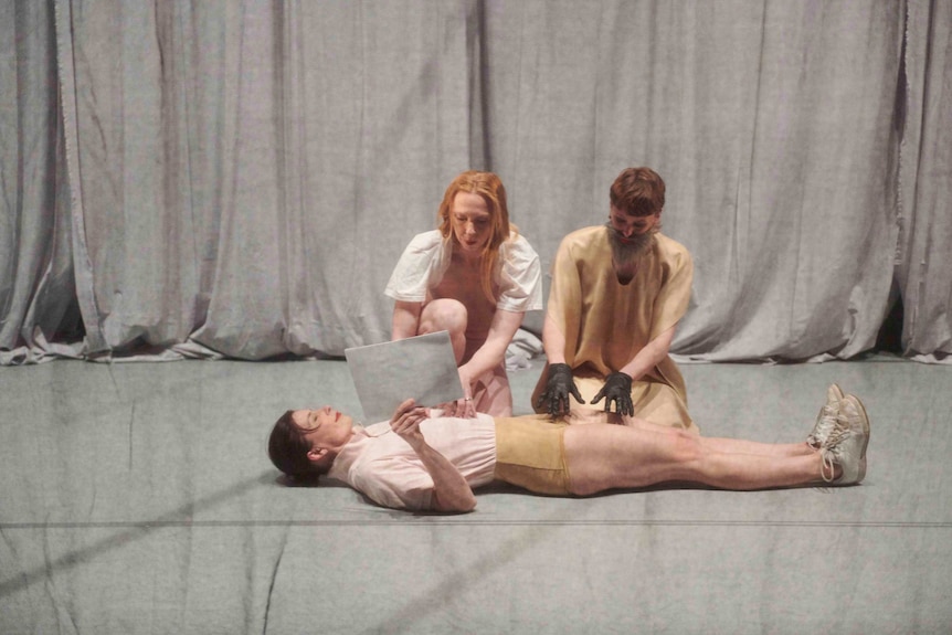 On a stage behind mesh material three dancers, one lies on the ground holding a piece of paper and the others touch her body
