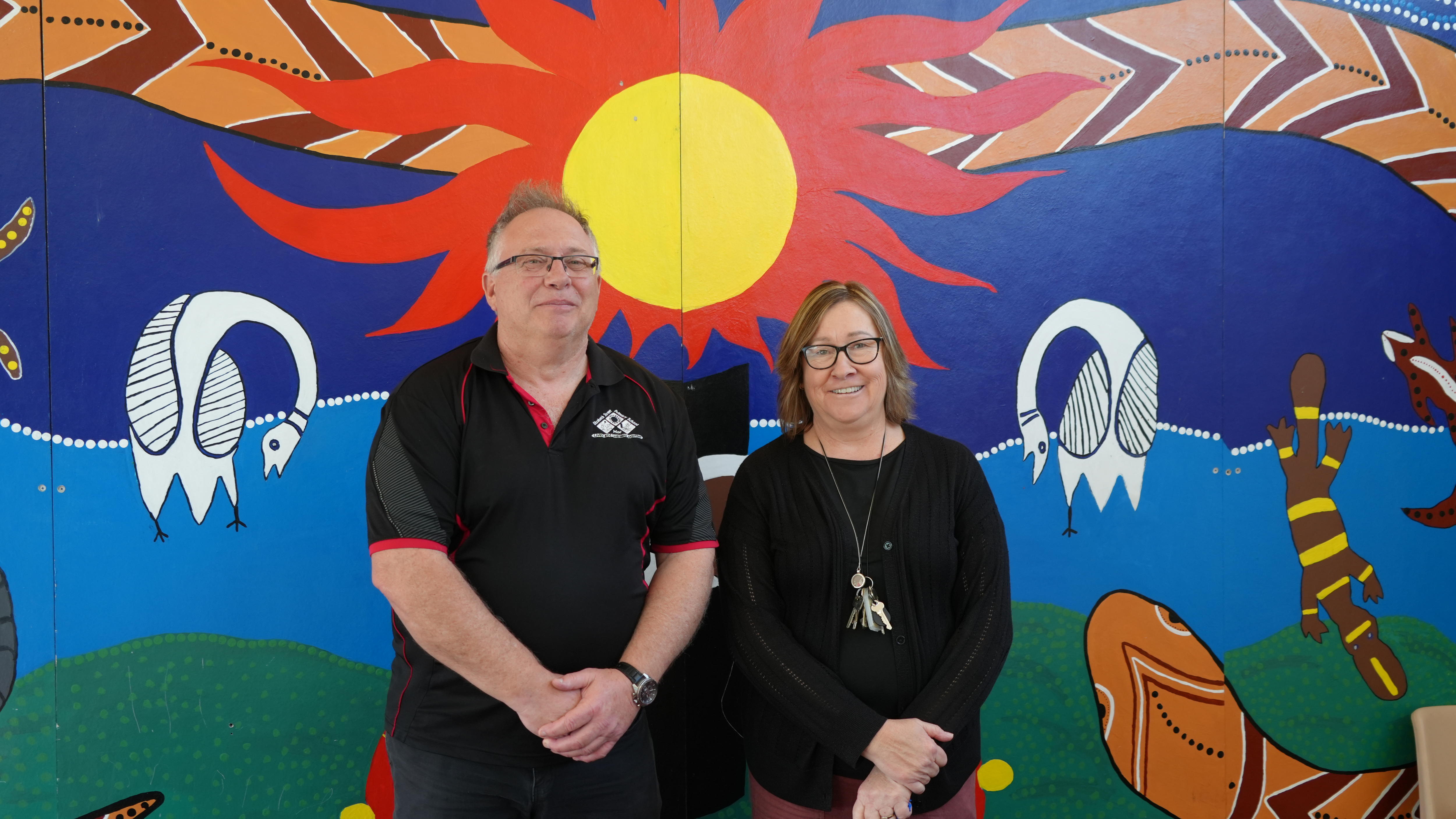 Ian Frost, with white hair, and Carol, with brown hair, stand in front of a painted sun mural.