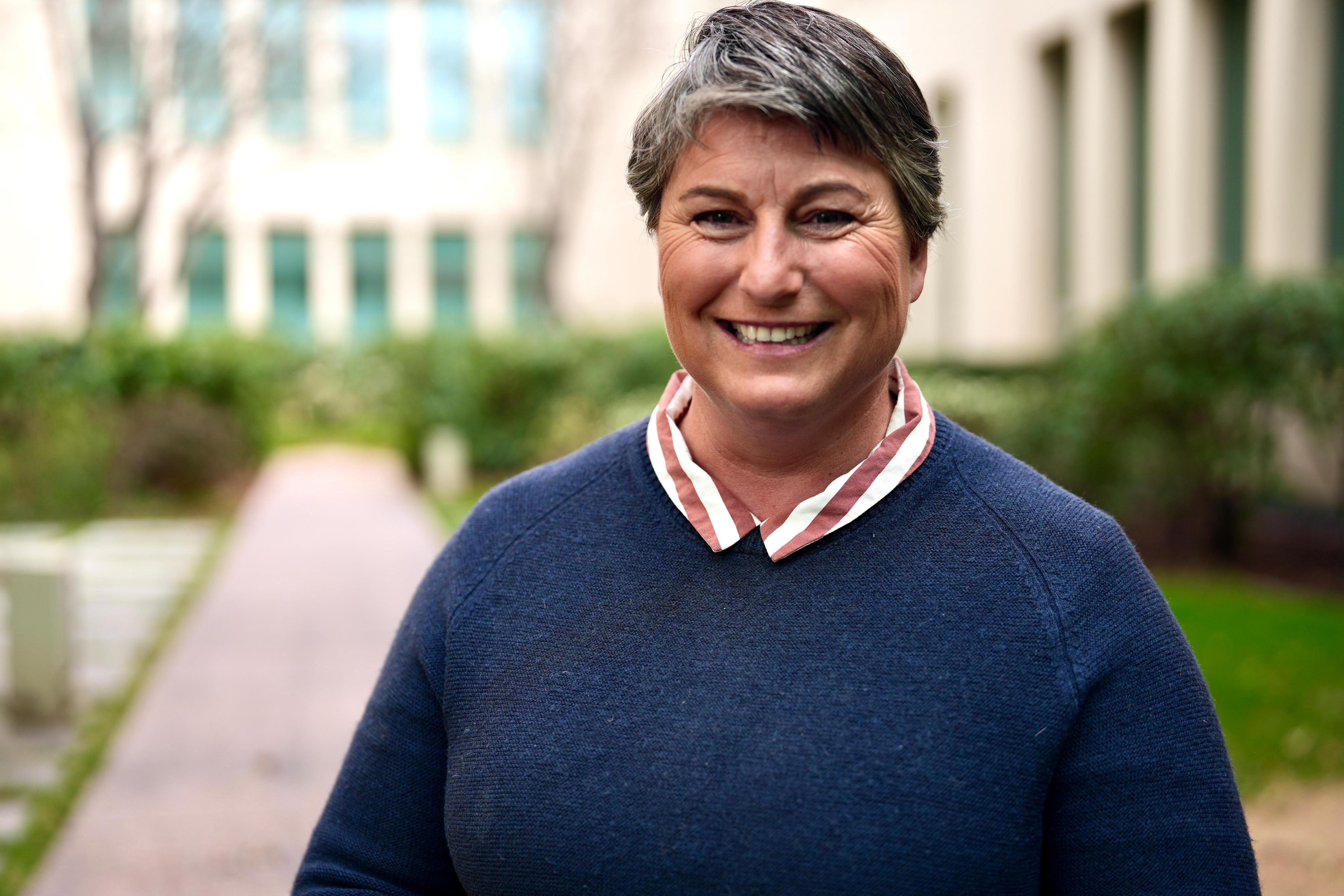 A woman with short hair smiles. She wears a dark blue jumper and a shirt with a striped collar.