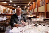 An older man sitting in his winery with a glass of wine in hand.