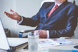 A man wearing a business suit gestures while sitting at a desk.