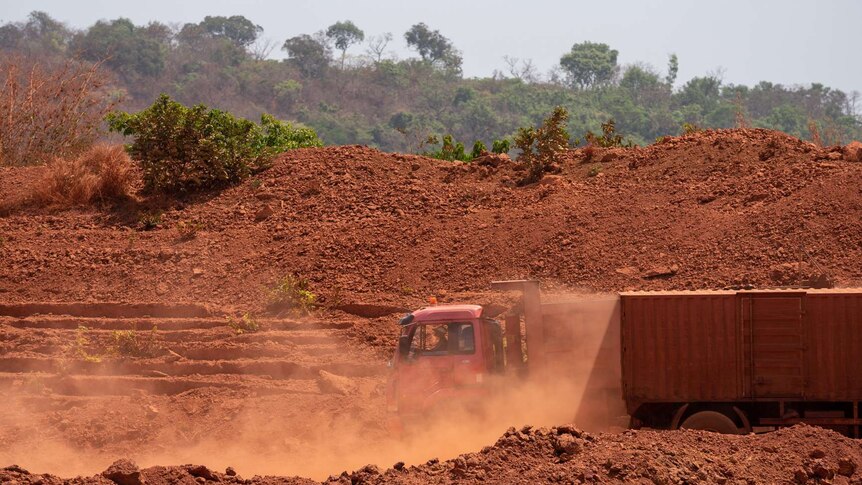 A truck drives down a brown dusty path into a mine cutting, with trees in background on a hill
