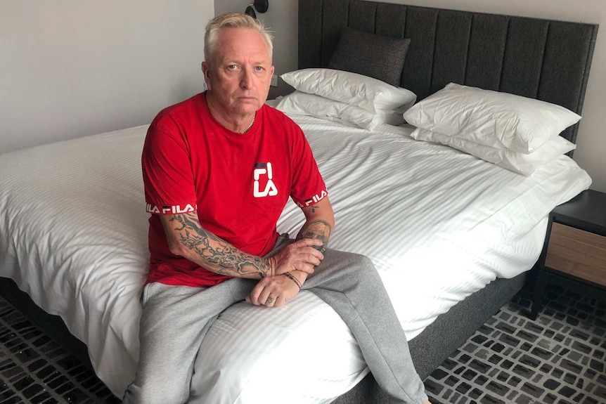 A man in a red t-shirt and grey tracksuit pants sits on a bed in a hotel room rooking at the camera.