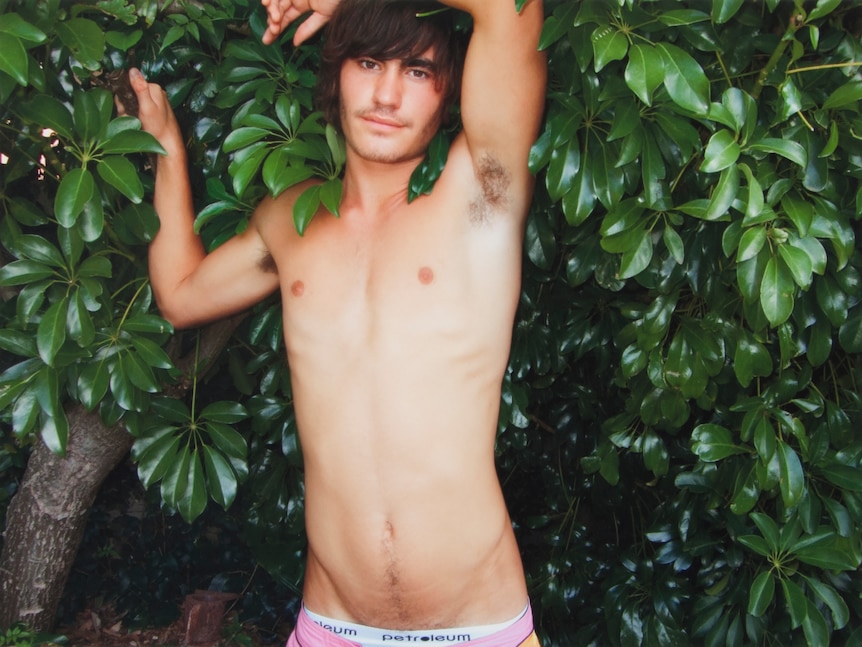 A man without a shirt on stands next to a green bush.