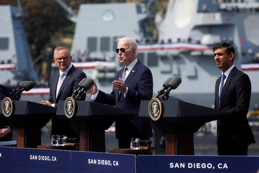 Anthony Albanese, Joe Biden, and Rishi Sunak stand behind lecterns outdoors. Behind them is a US Navy vessel.