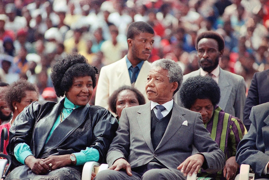 Winnie Mandela with her former husband Nelson Mandela. They are seated with a large crowd behind them.