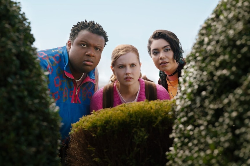 Teenage boy and two teenage girls stand behind a hedge.