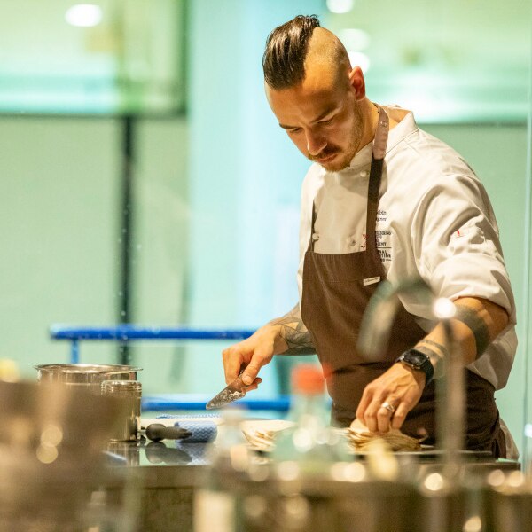 Chef with mohawk in white chef uniform, brown apron, knife in hand preparing a meal