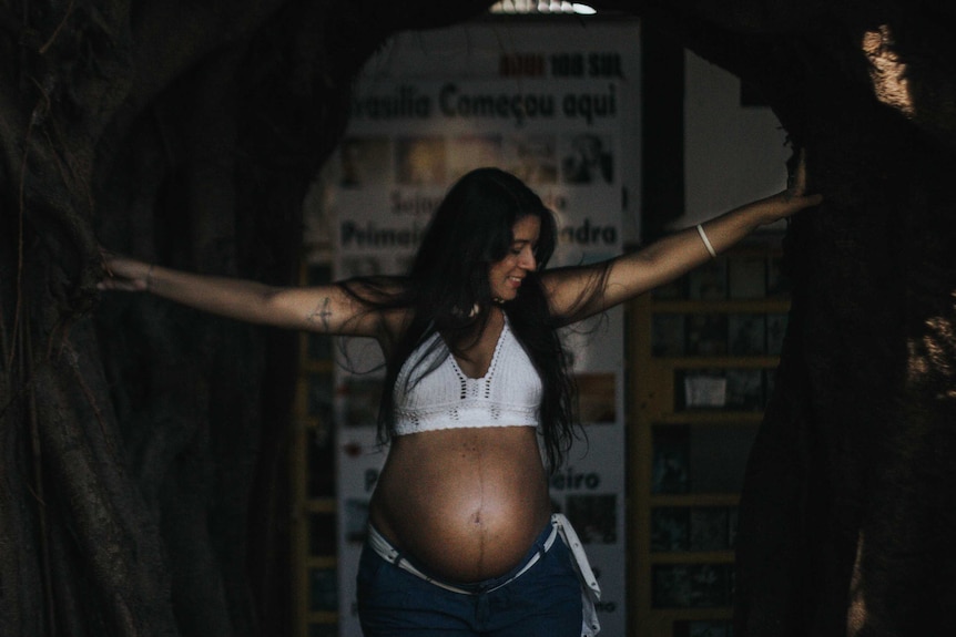 A pregnant woman with bare belly smiles and stands with arms wide in a doorway.