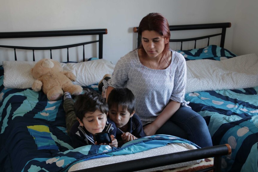 Hayfa Adi sits on a bed beside her two sons who are looking at a mobile phone screen.