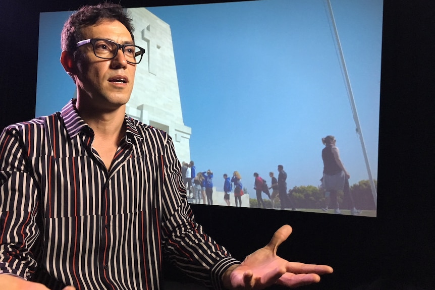 Artist Koken Ergun sits in front of a screen that is showing a scene from his film.