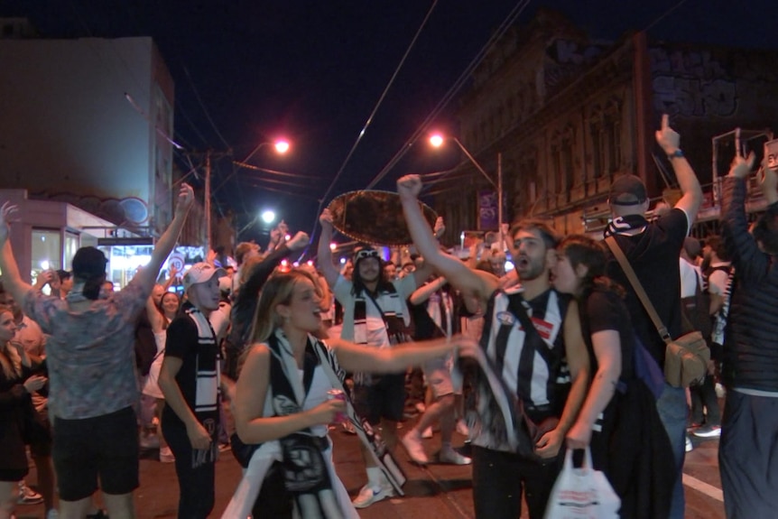 Fans wearing Collingwood guernseys cheer and throw their arms in the air in the middle of the street.