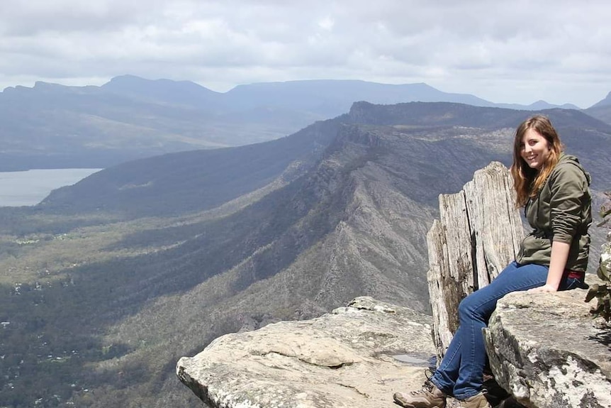 Woman in green jumper sits on giant rock, mountains in background.