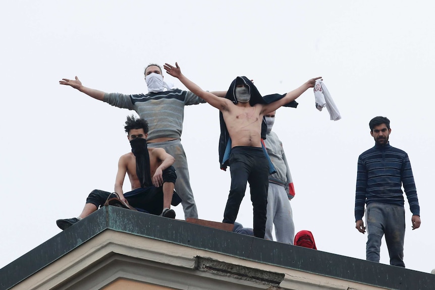 Young men, some wearing masks and some bare chested, wave from top of roof of building.