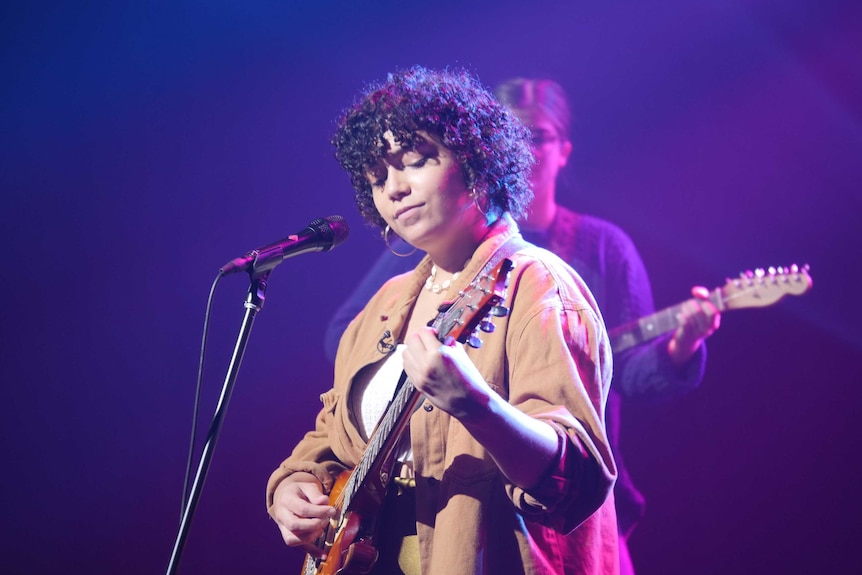 Musician Kee'ahn performs holding a guitar in front of a microphone on a purple-lit stage.