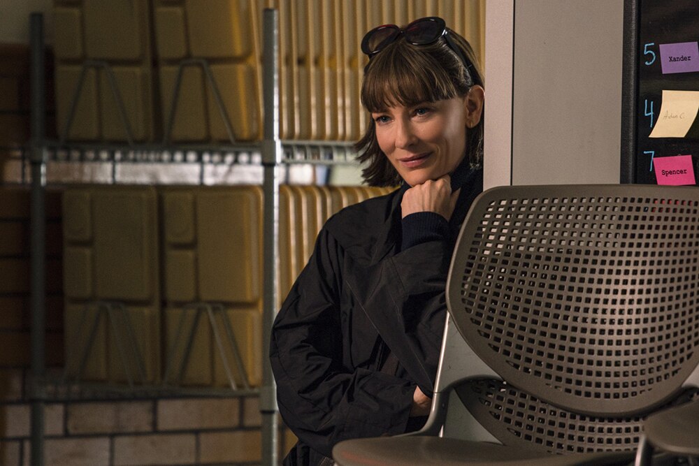 A woman with dark outfit, hair and sunglasses on head stands leaning against wall near corridor of stacked lunch trays.