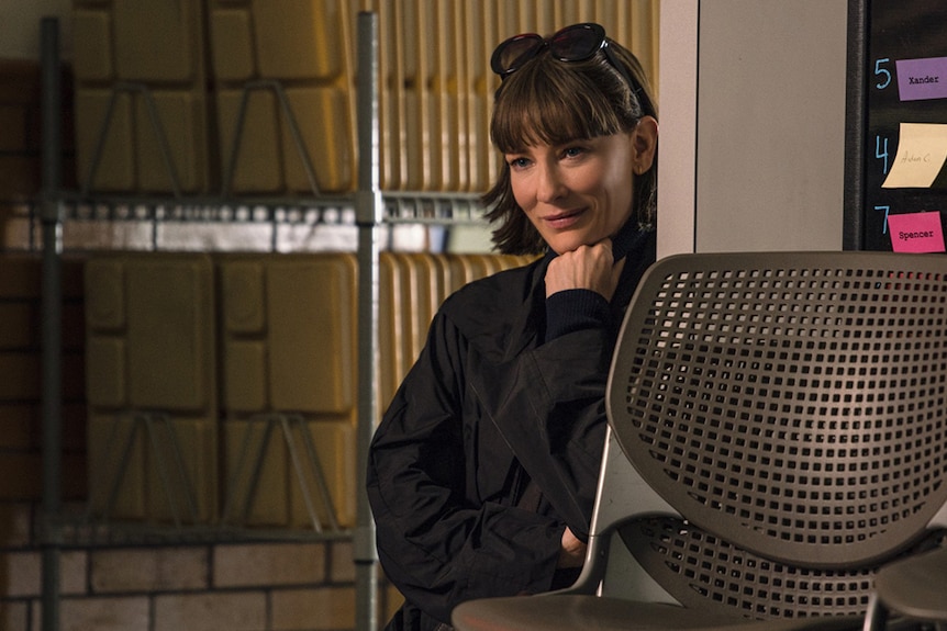 A woman with dark outfit, hair and sunglasses on head stands leaning against wall near corridor of stacked lunch trays.