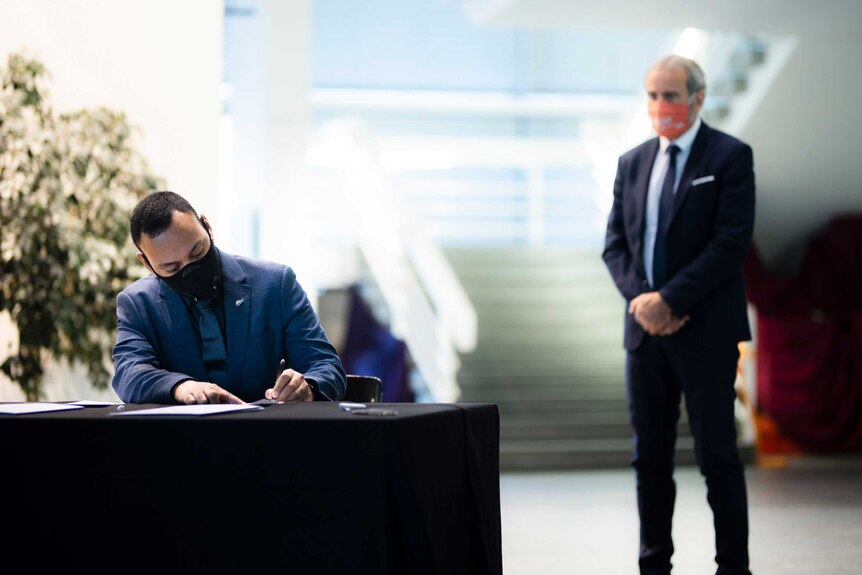 A man in a suit and face-mask sits at a table, signing a formal document as another man watches on.