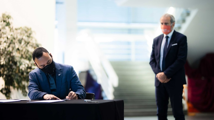 A man in a suit and face-mask sits at a table, signing a formal document as another man watches on.