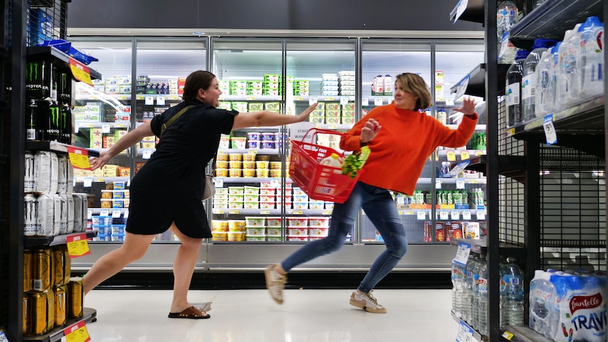A woman in a red jumper and blue jeans, holding a basket, tries to avoid being 'tagged' by another woman in a supermarket.