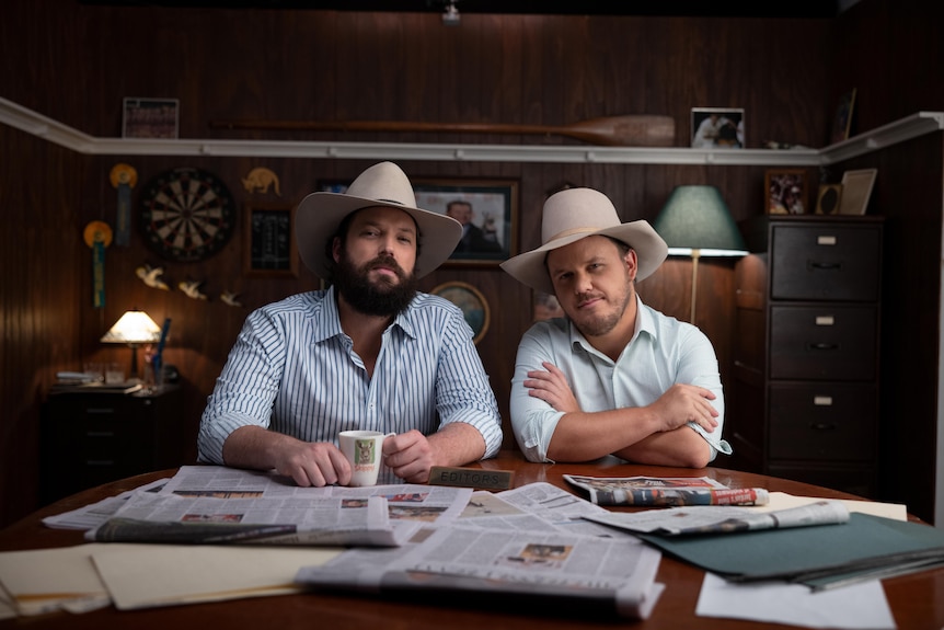 Clancy Overell and Errol Parker sitting at a desk with newspapers in front of them, one holding a coffee mug, both in Akubras