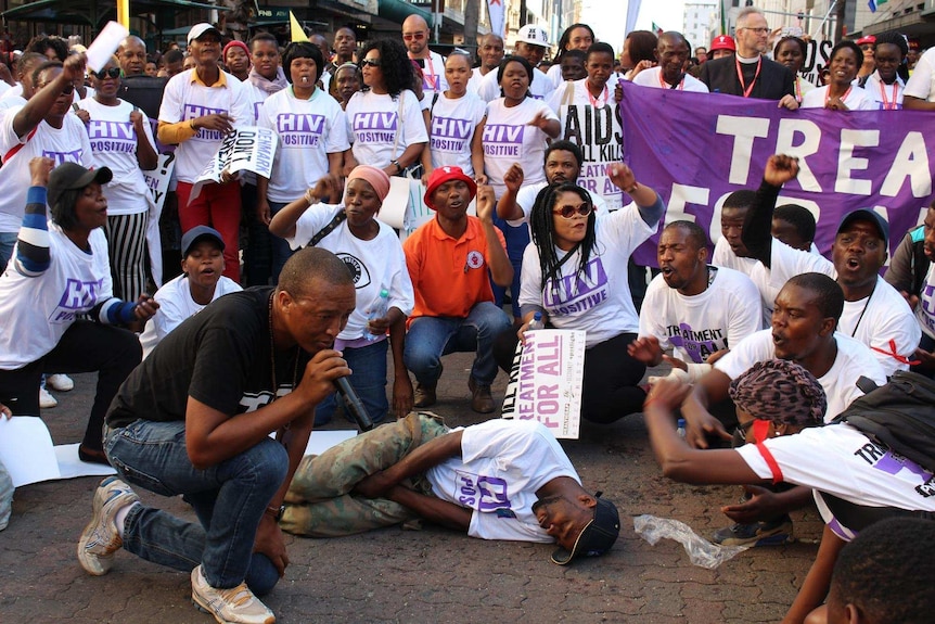 An activist falls to the ground next to a man with a microphone, surrounded by activists wearing t-shirts saying 'HIV-positive'.