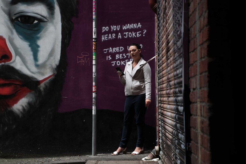 A picture of a woman holding a phone looking at the camera. Graffiti art on the wall behind shows a clown.