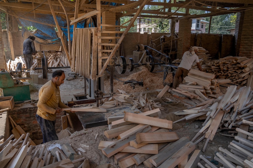 Trois hommes se tiennent à des postes de travail individuels au milieu de tas de battes de cricket en bois dans un atelier.