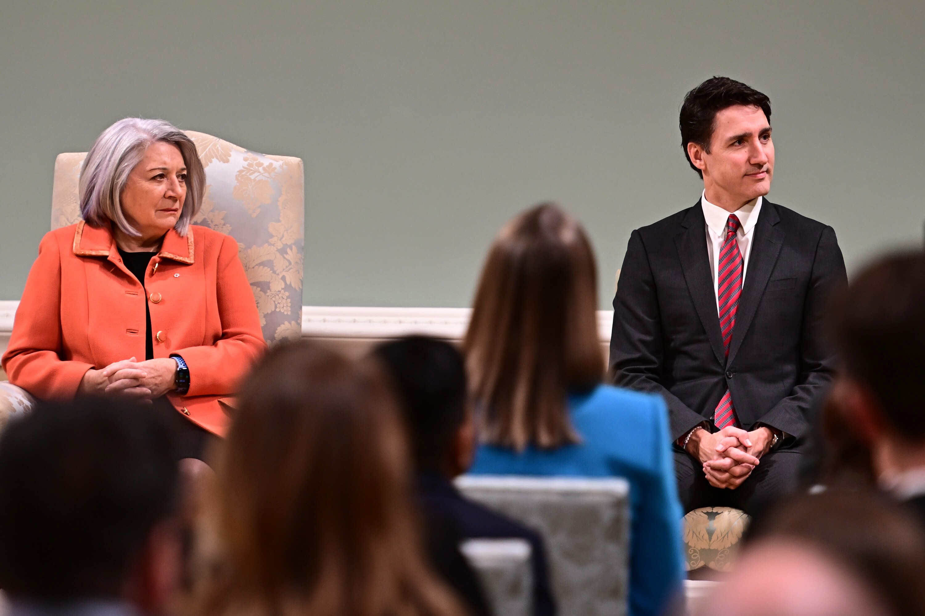A woman in an orange blazer sits to the right of a man in black suit and red tie