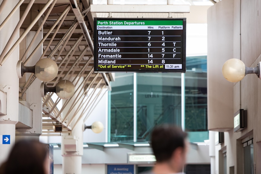 A TV screen showing train departure information, hanging from a roof with two people in the foreground.